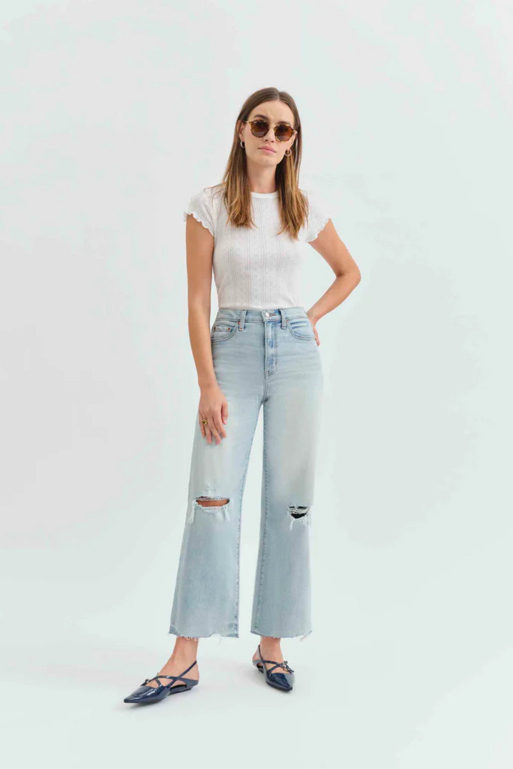 Front view of a woman wearing a white t-shirt with light denim jeans with holes in the knees, and pointed flats. 