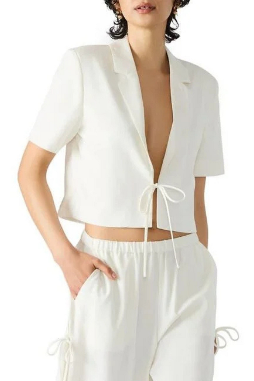 A woman wearing a white cropped blazer with a tie in front, and matching white pants.