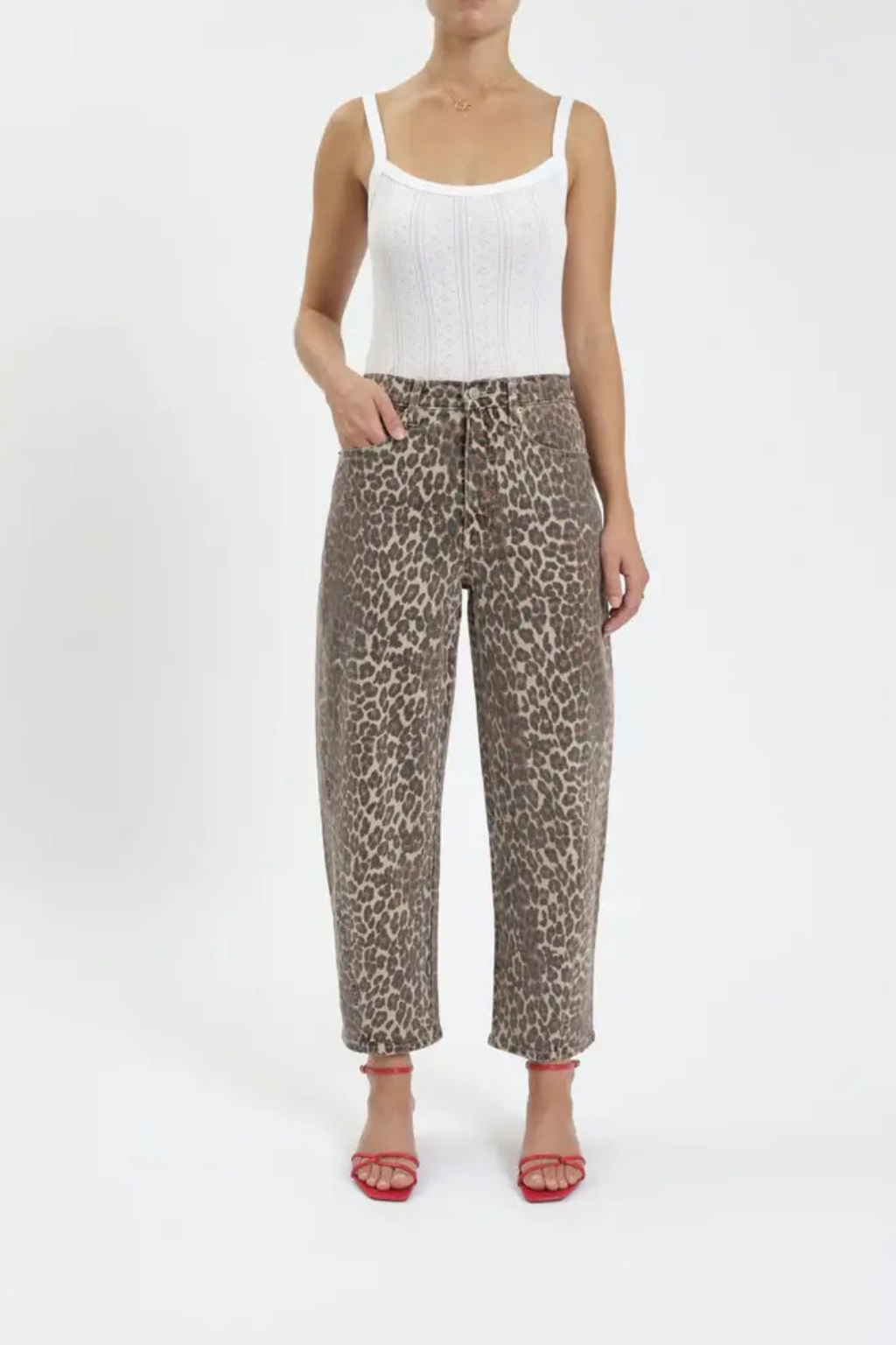 A front view of a woman wearing a white tank top with leopard barrel leg jeans, and red heels. 