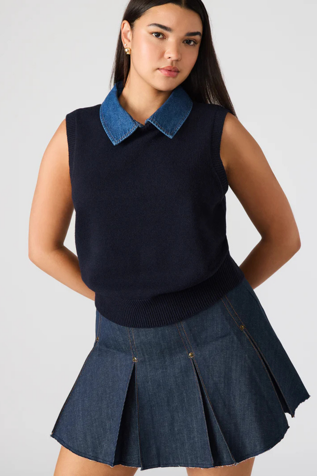 Woman wearing a navy sleeveless top with a blue collar and pleated denim skirt on a white background.