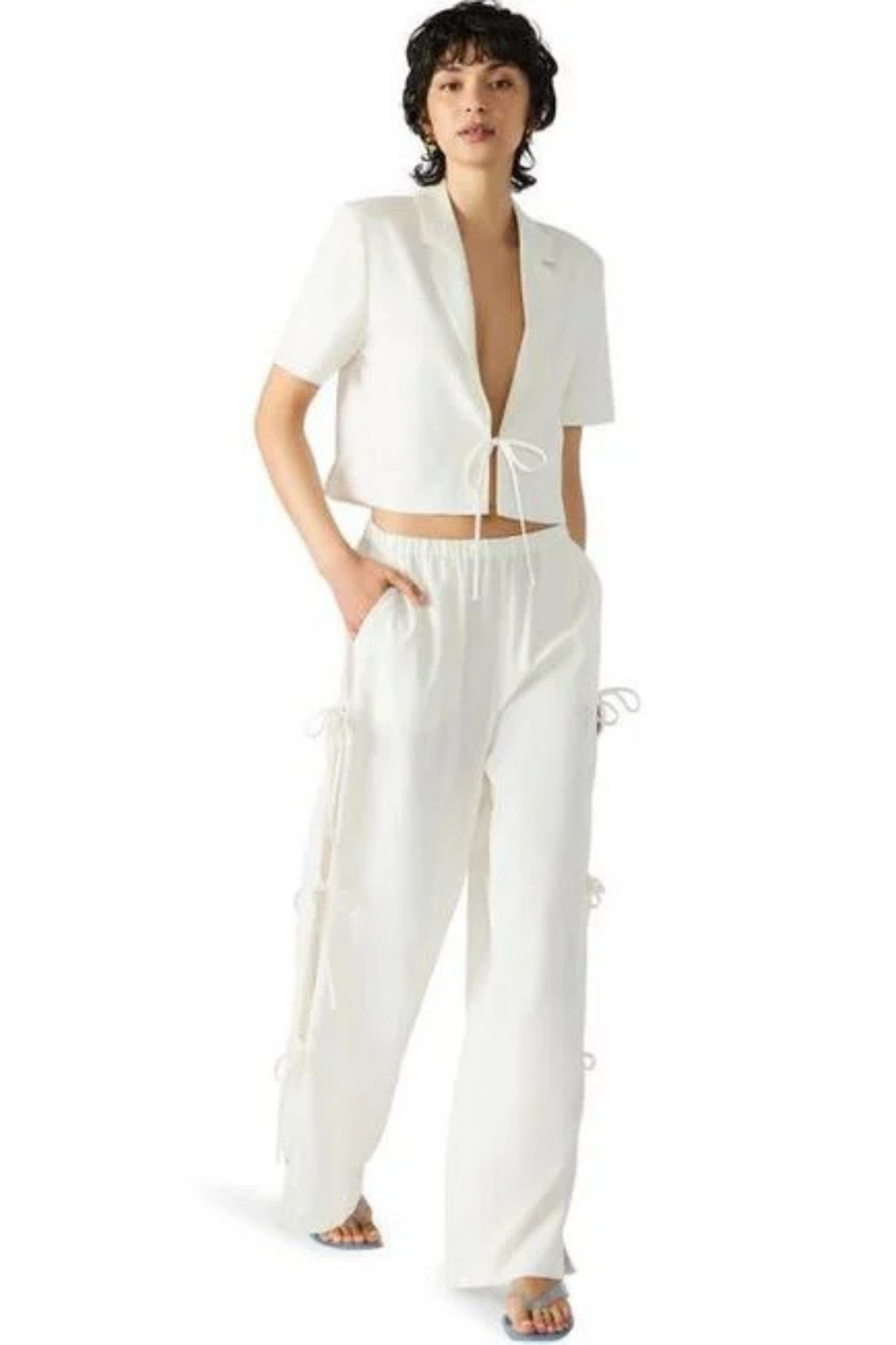 A woman wearing a white cropped blazer with a tie in front, and matching pants.