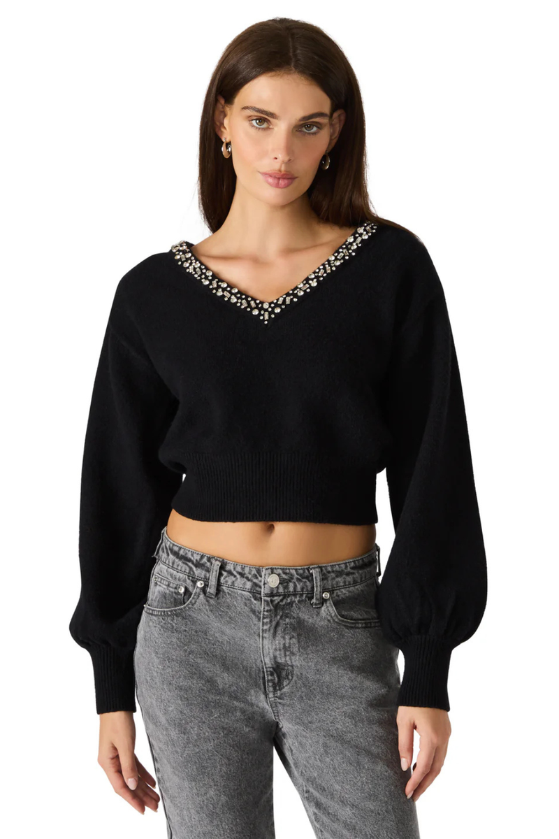 Woman wearing a black v neck sweater with rhinestones on the neckline and faded grey jeans. 
