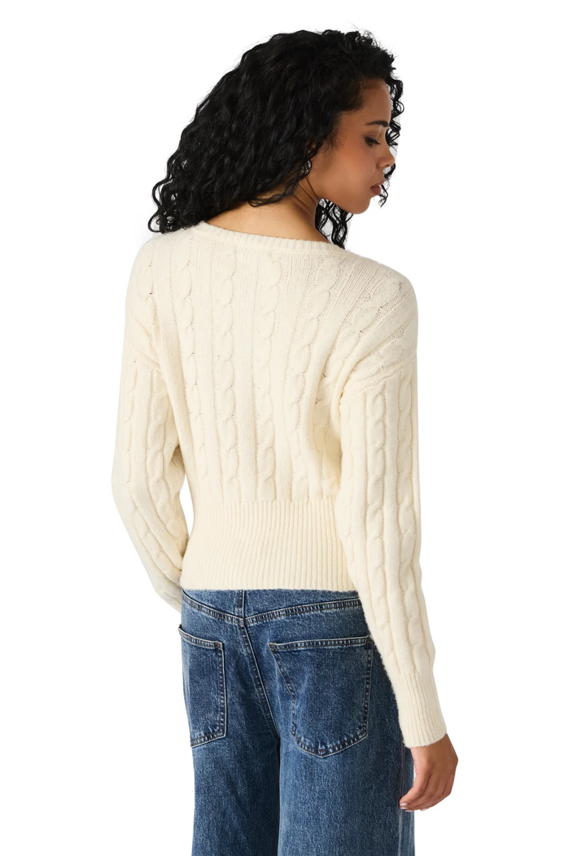 Woman wearing a cream cable knit sweater and blue jeans on a white background.