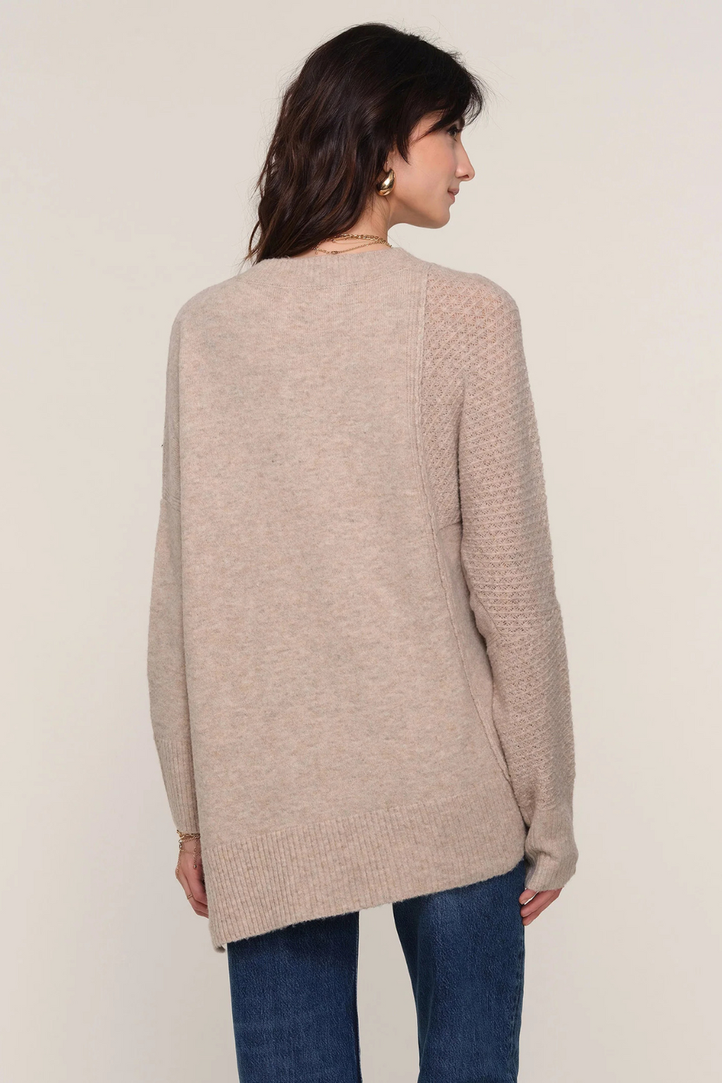 Woman wearing a beige sweater and blue jeans on a plain background.