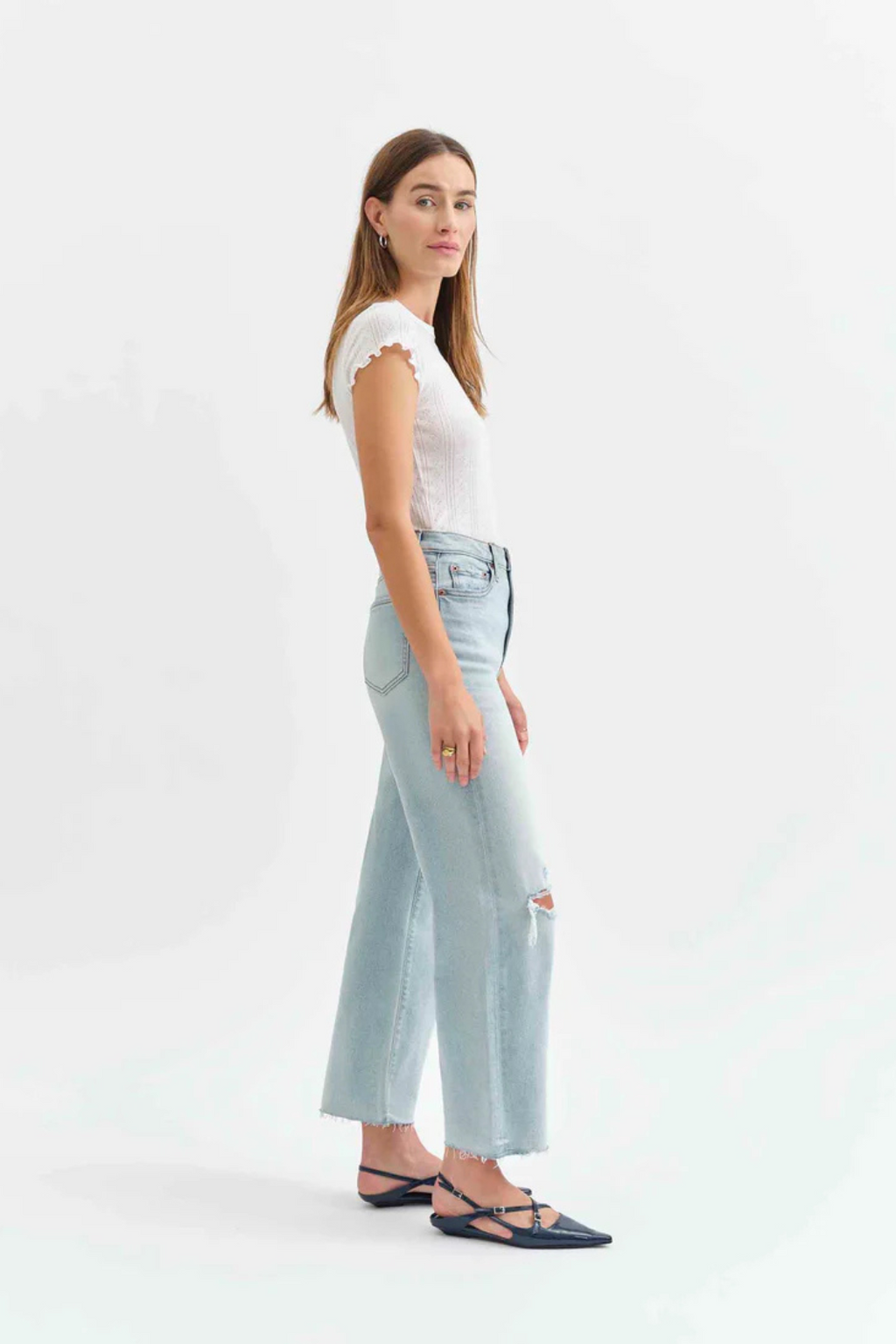 Side view of a woman wearing a white t-shirt with light denim jeans with holes in the knees, and pointed flats. 