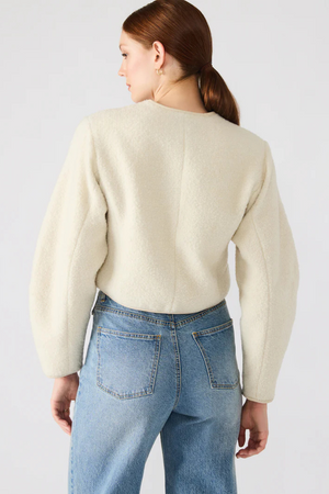 Person wearing a beige jacket and blue jeans on a plain background.