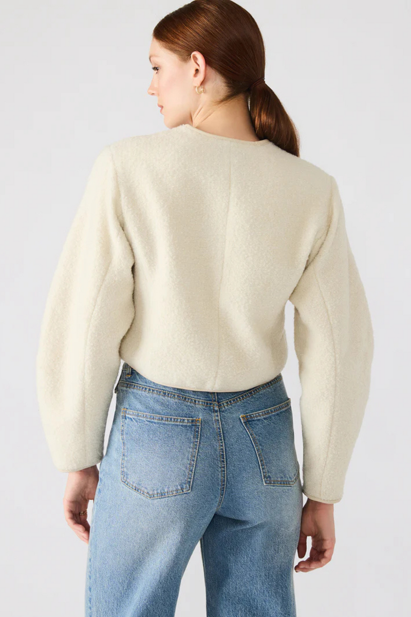 Person wearing a beige jacket and blue jeans on a plain background.