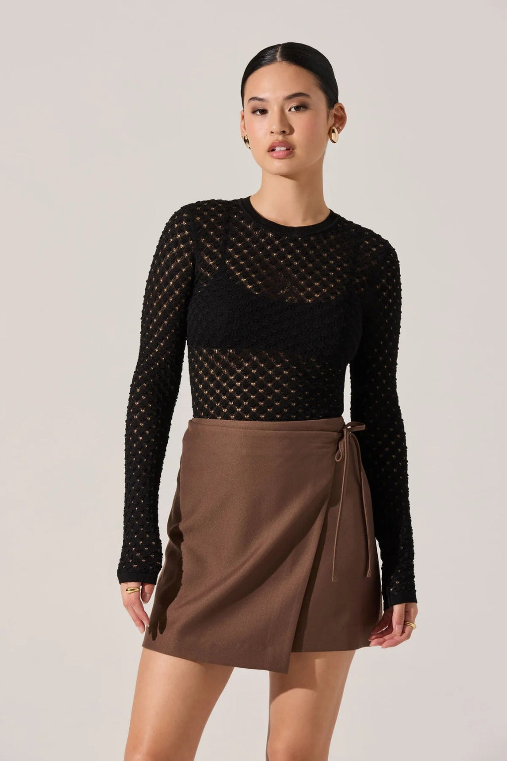 Model wearing a black mesh top and brown wrap skirt on a plain background.