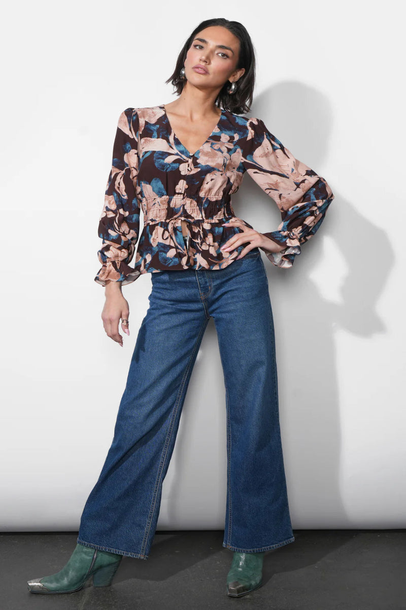 Woman wearing a patterned blouse and blue jeans against a white background.