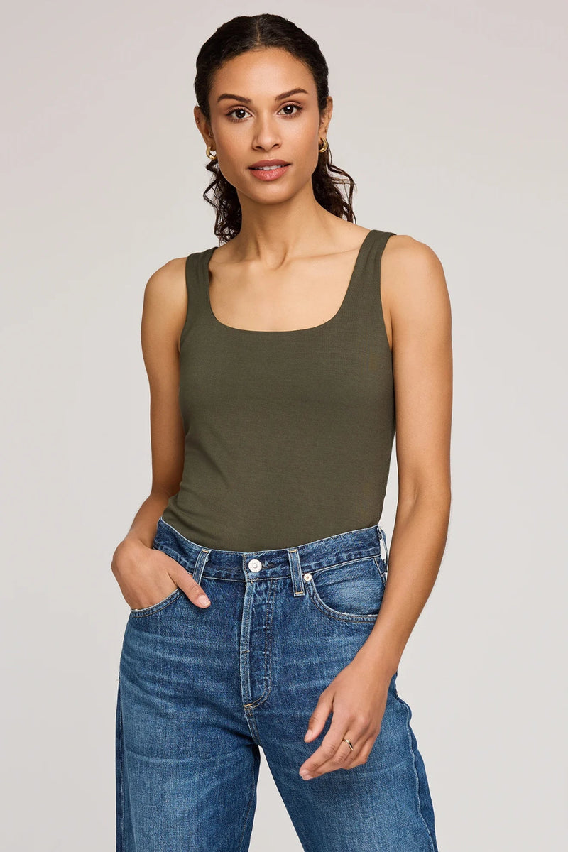 Woman wearing an olive green tank top and blue jeans on a plain background.
