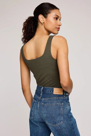 Woman wearing an olive green tank top and blue jeans on a plain background.