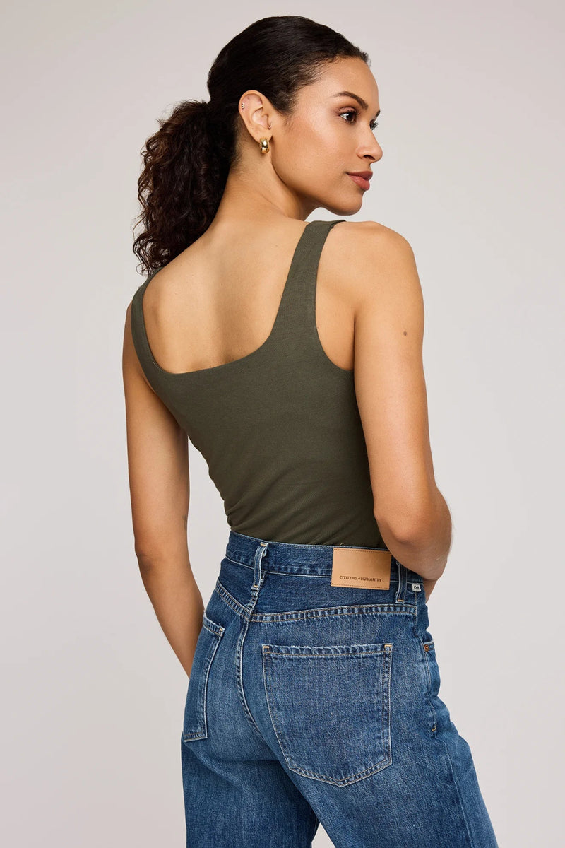Woman wearing an olive green tank top and blue jeans on a plain background.