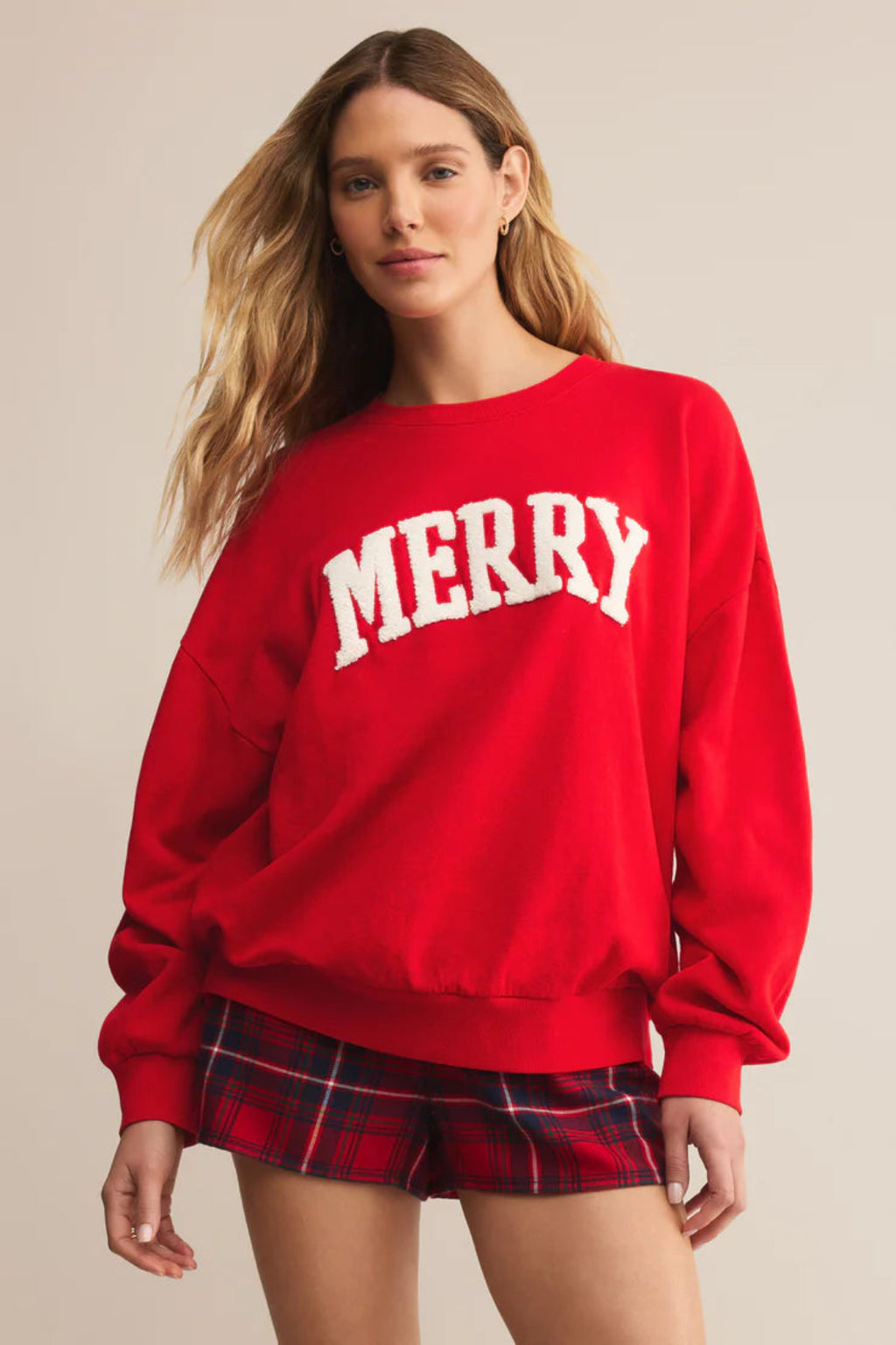 Person wearing a red 'MERRY' sweatshirt and plaid shorts on a beige background.