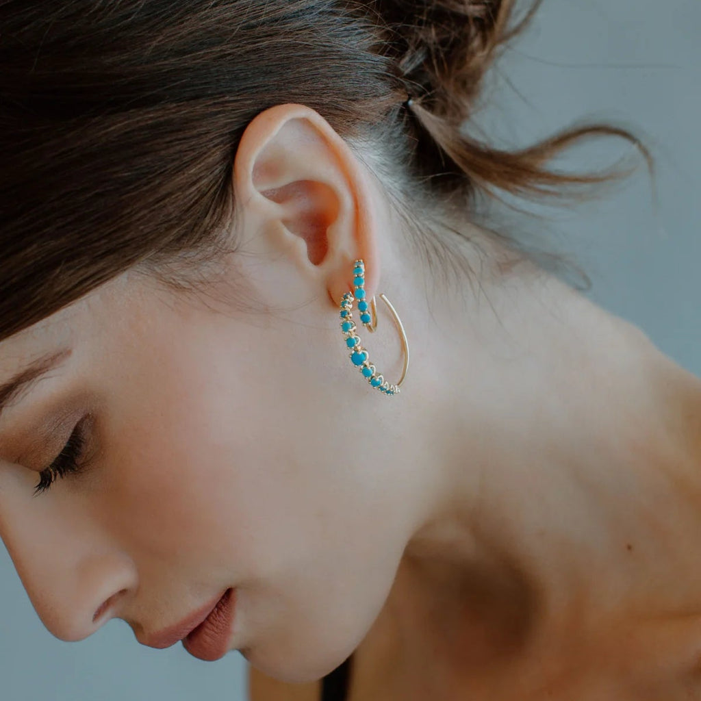 Close-up of a person wearing gold hoop earrings with blue beads on a neutral background