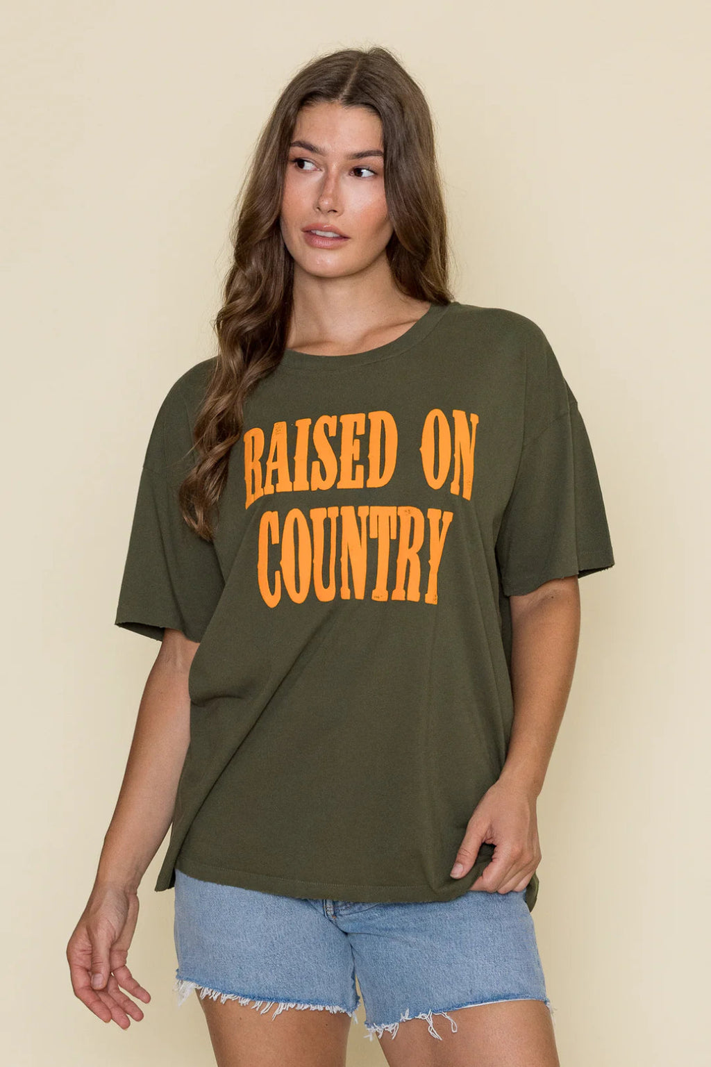 Straight on view of a woman wearing a green shirt with orange writing, and denim shorts. 
