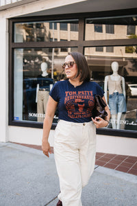 Woman wearing a 'Tom Petty' t-shirt walking on a sidewalk with a store window in the background.
