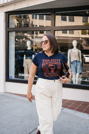 Woman wearing a 'Tom Petty' t-shirt walking on a sidewalk with a store window in the background.