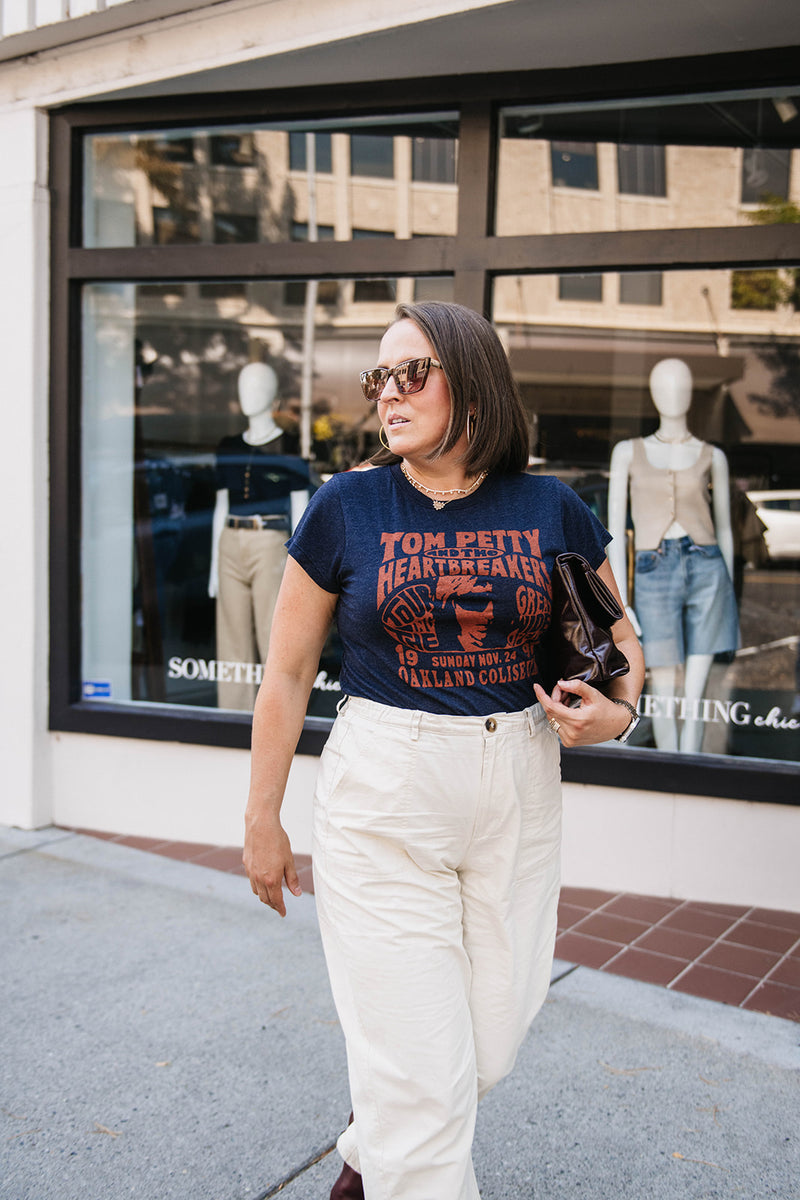Woman wearing a 'Tom Petty' t-shirt walking on a sidewalk with a store window in the background.