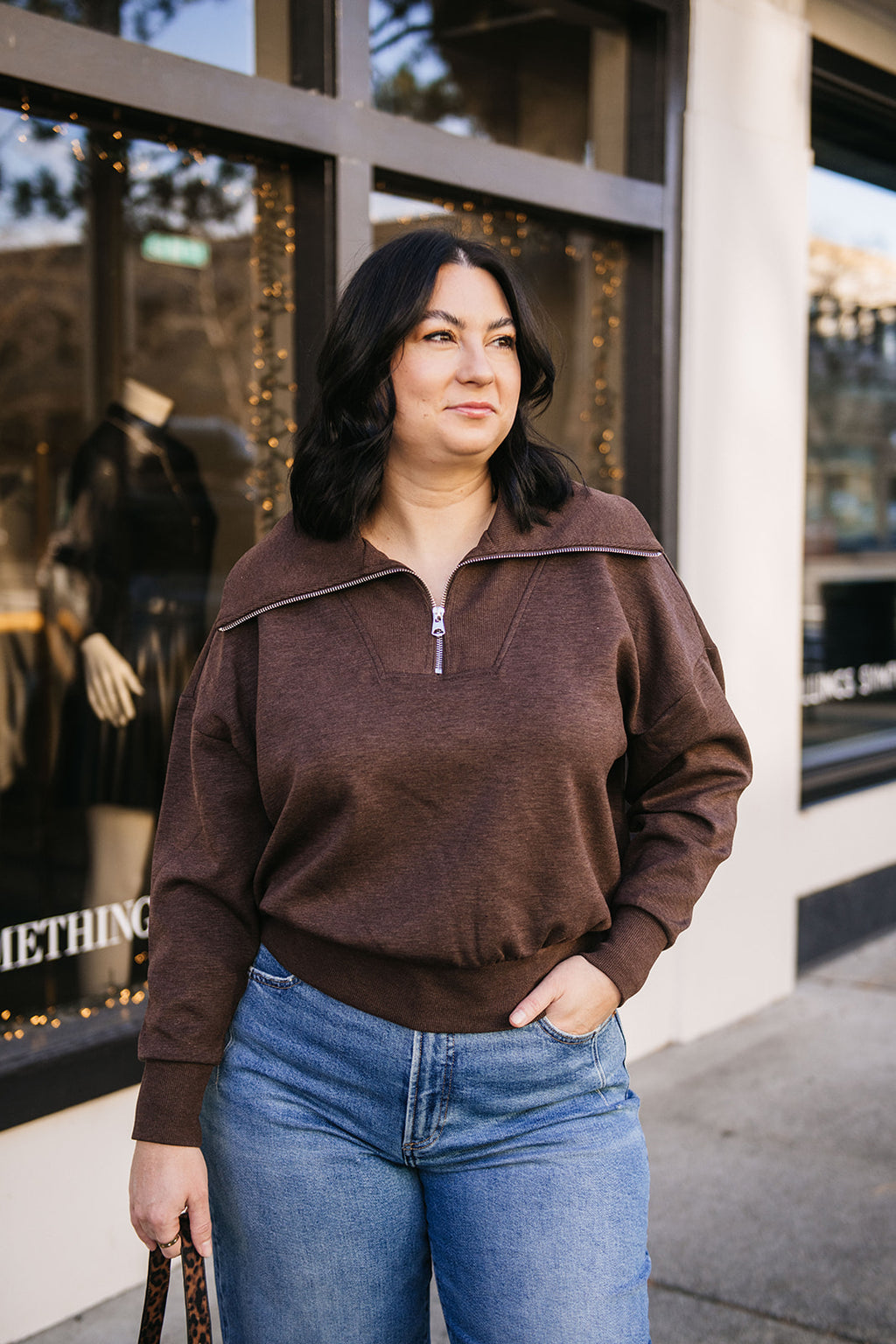Woman wearing a brown sweatshirt and blue jeans standing in front of a store window.