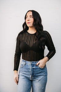 Woman wearing a black long-sleeve top with sheer stripes and high-waisted blue jeans against a plain background.