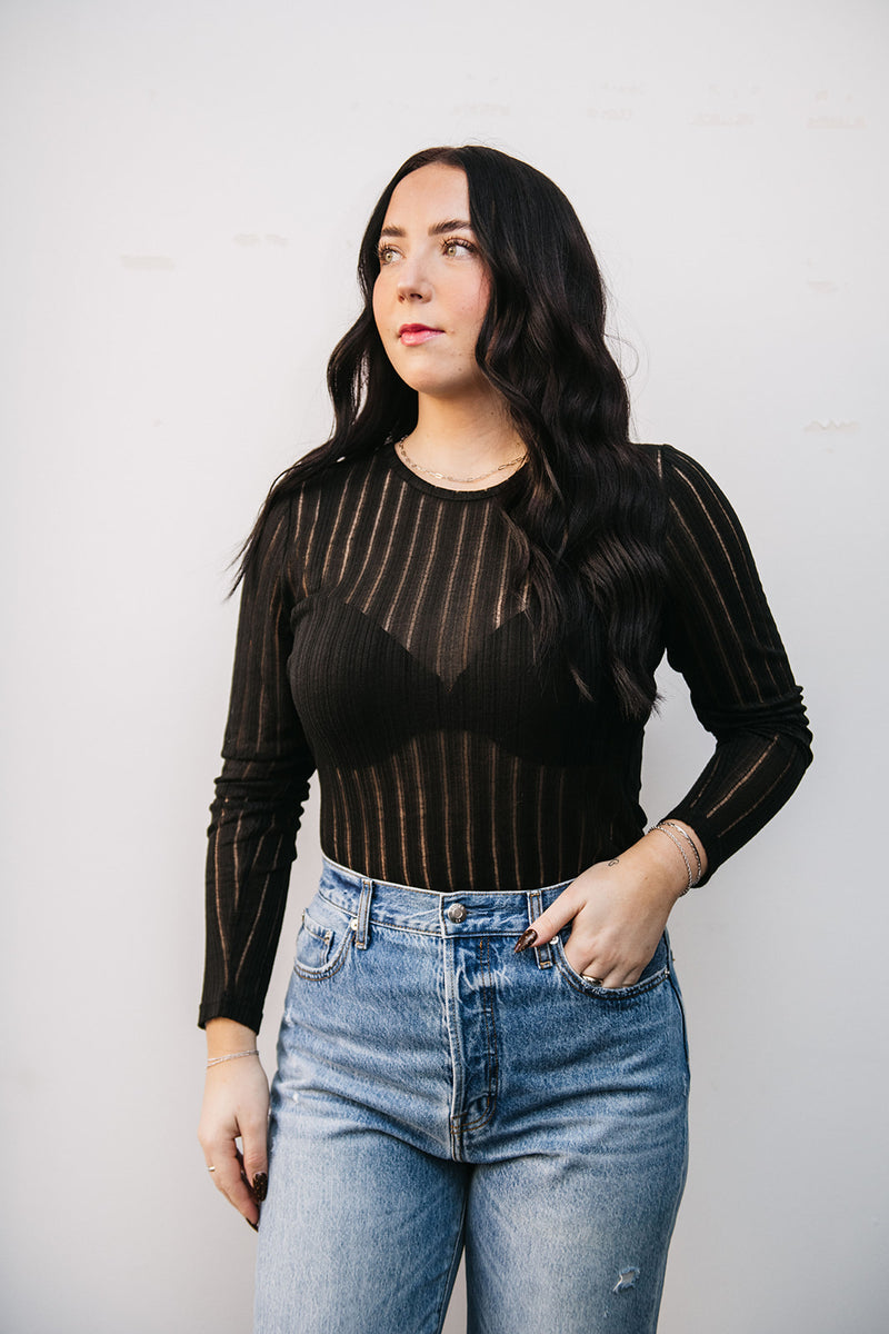 Woman wearing a black long-sleeve top with sheer stripes and high-waisted blue jeans against a plain background.