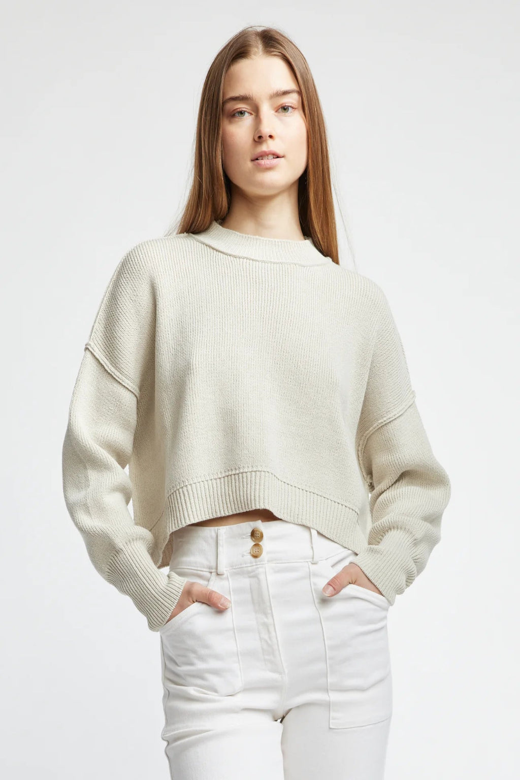 Woman wearing a beige sweater and white pants on a plain background.