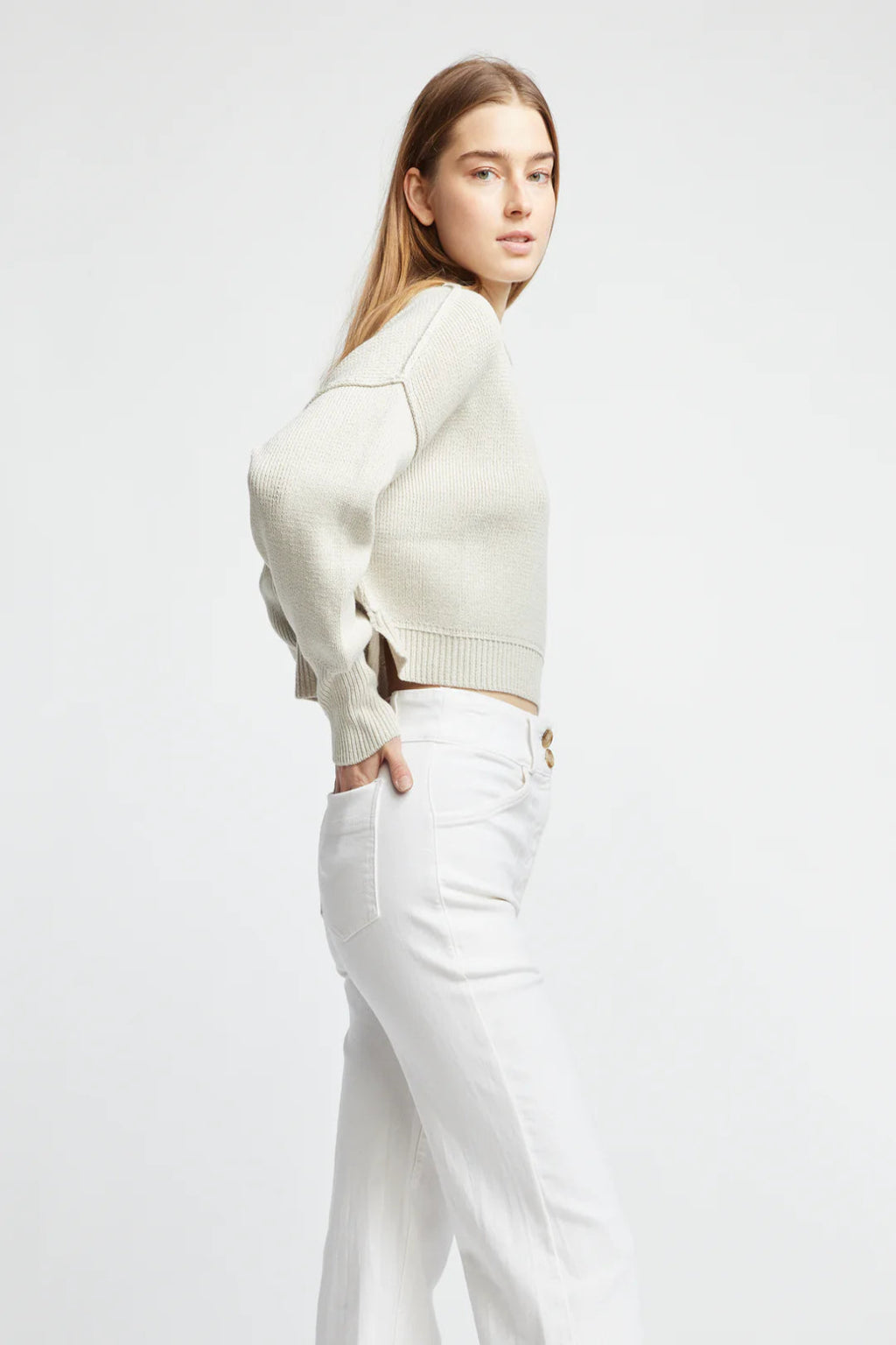 Woman wearing a cream sweater and white pants on a light gray background.