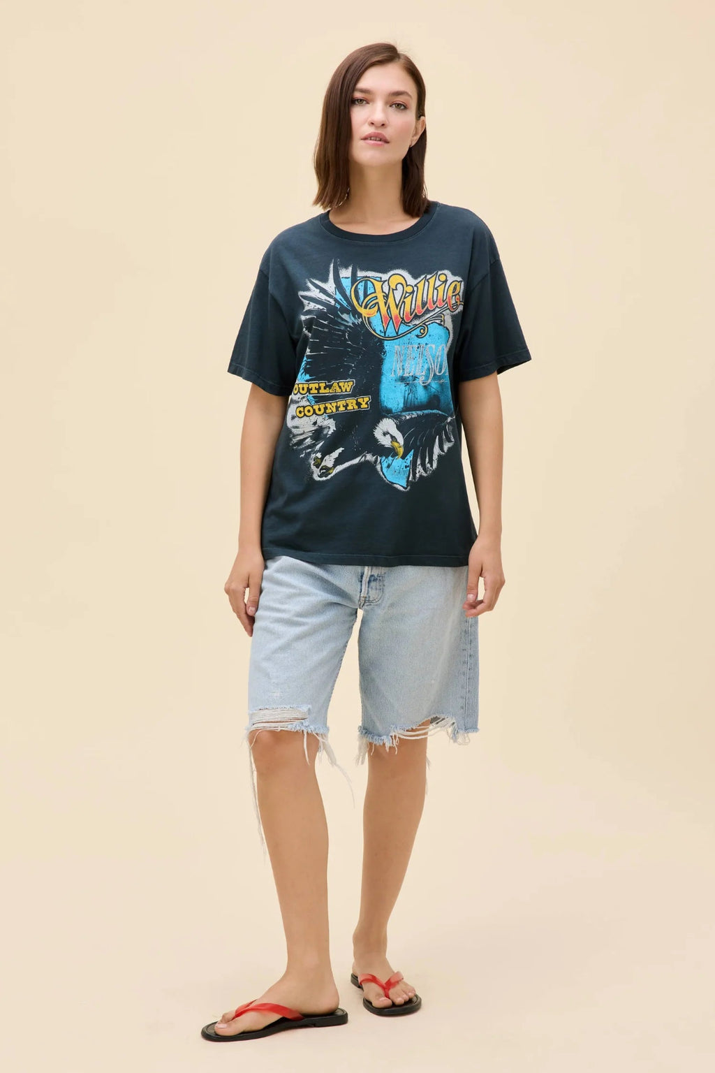 Straight on view of a woman wearing a navy blue Willie Nelson graphic tee with denim shorts and red & black sandals.