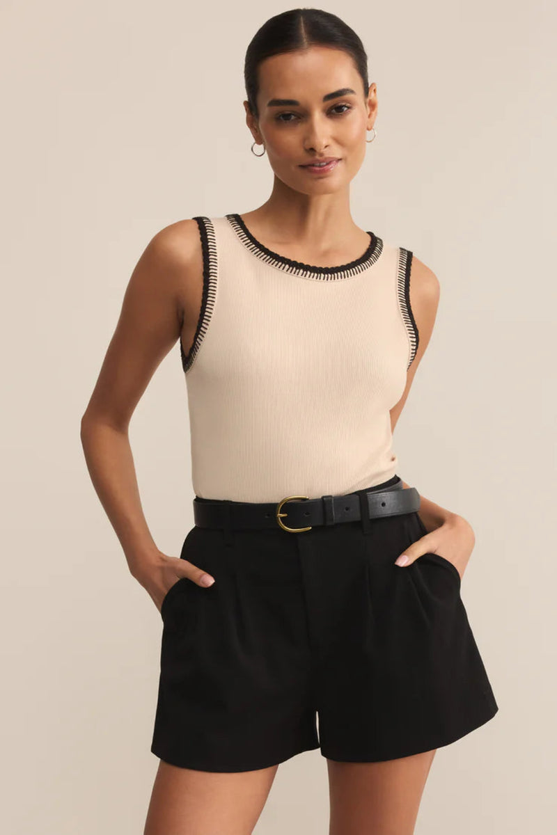Woman wearing a beige sleeveless top with black trim and black shorts on a beige background.