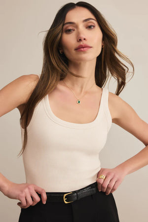 Woman wearing a beige tank top and black pants against a plain background.