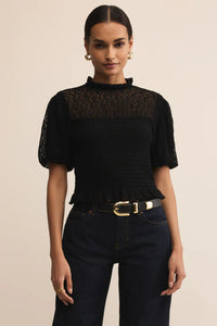Woman wearing a black lace top and dark jeans against a beige background.
