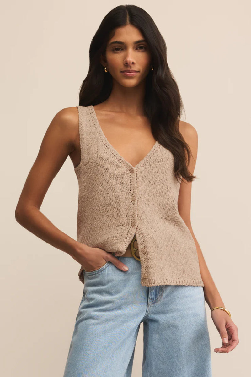 Woman wearing a beige knitted vest and light blue jeans against a beige background.