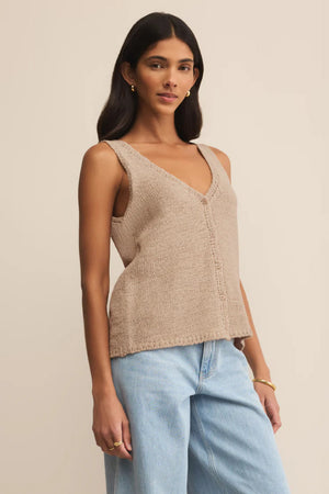 Woman wearing a beige knitted vest and light blue jeans against a beige background.