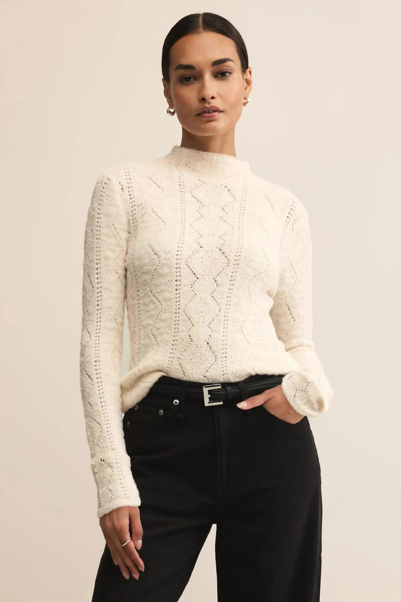 Woman wearing a cream cable knit sweater and black pants against a beige background.
